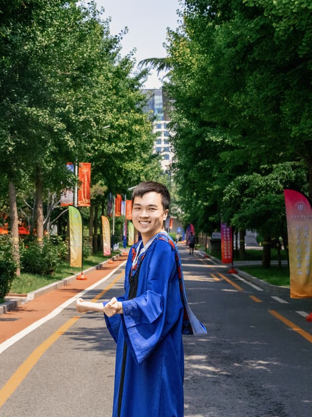 一位身穿蓝色硕士服的年轻男子站在绿树成荫的道路上,面带微笑回望镜头,道路两侧立有彩色道旗。